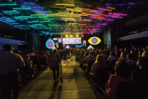 Photo of the inside of the convention center. Dark room with people seated and colorful lights on the ceiling 